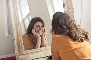 Consultoria de Imagem e Estilo - Mulher bonita na frente de um espelho, feliz e sorrindo ao olhar para a própria imagem.